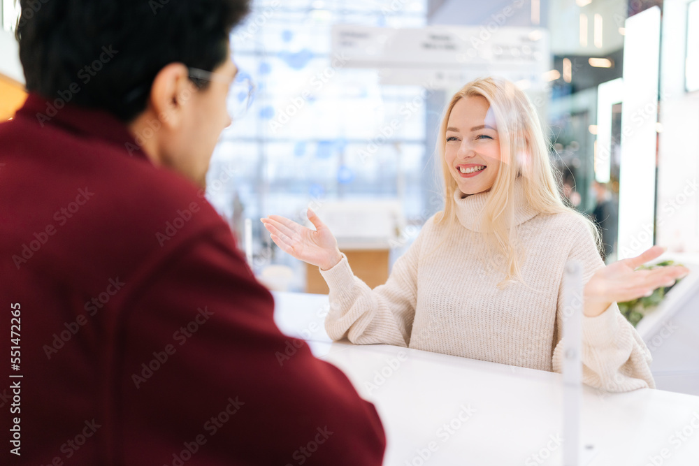 Back view from shoulder of unrecognizable man chatting through glass partition with cheerful woman standing at desk in bright hall of shopping mall. Concept of lifestyle social distancing and work. Back view from shoulder of unrecognizable man chatting through glass partition with cheerful woman standing at desk in bright hall of shopping mall. Concept of lifestyle social distancing and work.