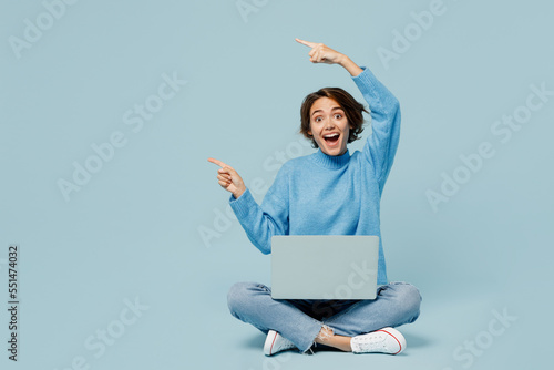 Full body young IT woman in knitted sweater hold use work on laptop pc computer point finger aside on area isolated on plain pastel light blue cyan background studio portrait People lifestyle concept.
