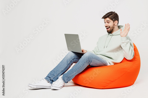 Full body young smiling fun IT man wears mint hoody sit in bag chair hold use work on laptop pc computer waving hand get video call isolated on plain solid white background People lifestyle concept.