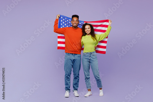 Full body fun young couple two friend family man woman of African American ethnicity wear casual clothes together hold American flag behind themselves isolated on pastel plain light purple background