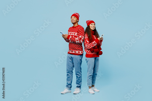 Full body minded merry young couple two man woman wear red Christmas sweater Santa hat posing hold use mobile cell phone isolated on plain pastel blue background. Happy New Year 2023 holiday concept.