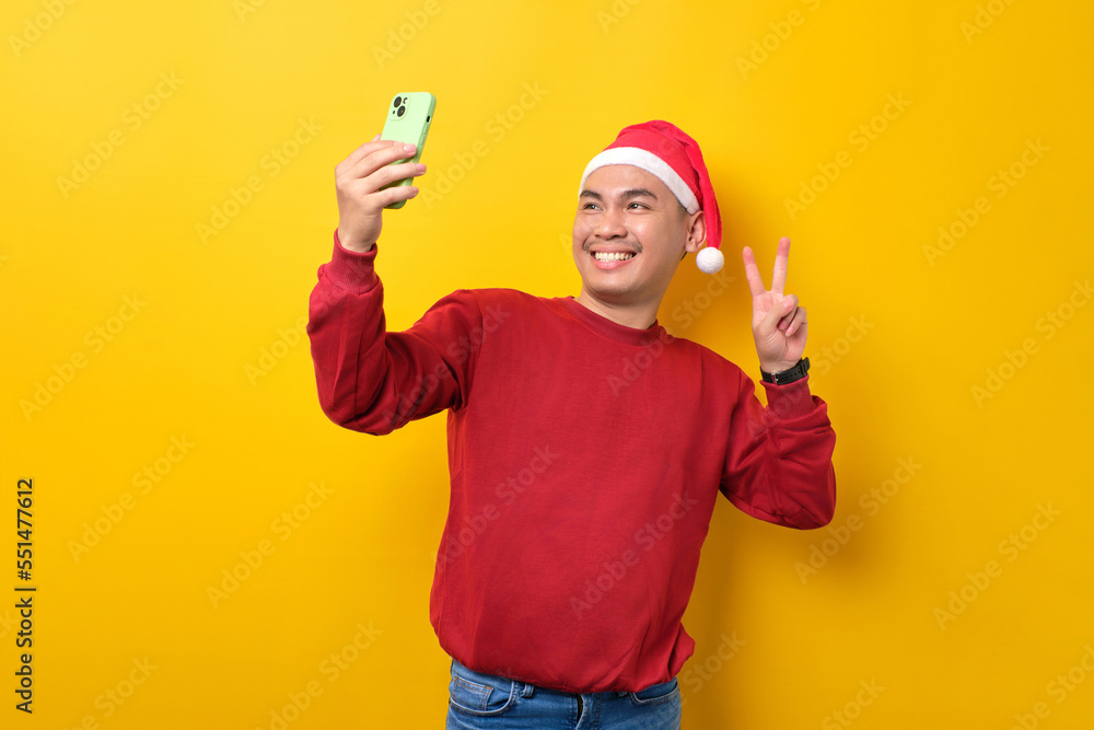 Cheerful young Asian man in Santa hat taking selfie on smartphone, showing victory sign over yellow studio background. celebration Christmas holiday and New Year concept Cheerful young Asian man in Santa hat taking selfie on smartphone, showing victory sign over yellow studio background. celebration Christmas holiday and New Year concept
