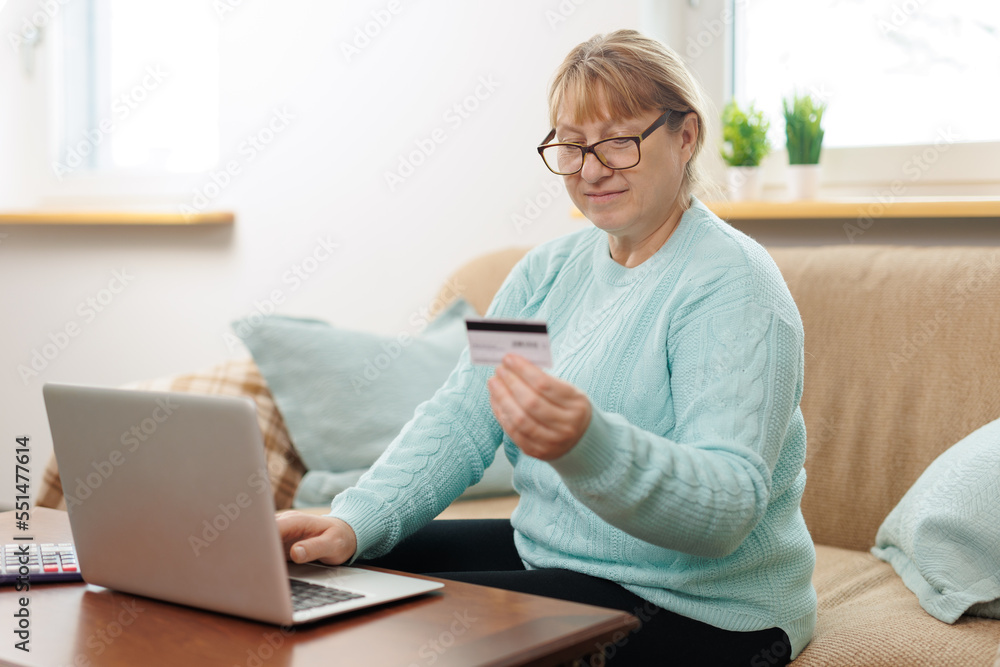 Clever senior satisfied woman sitting in the bright room on the sofa using the laptop and holding credit card. Clever senior satisfied woman sitting in the bright room on the sofa using the laptop and holding credit card.