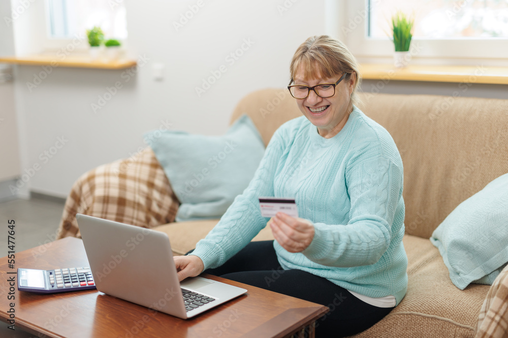Clever senior satisfied woman sitting in the bright room on the sofa using the laptop and holding credit card. Clever senior satisfied woman sitting in the bright room on the sofa using the laptop and holding credit card.