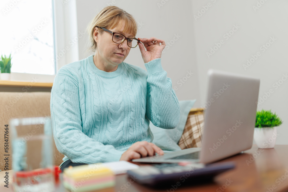 Happy mature businesswoman accountant in glasses and turquoise jumper sitting on the couch with laptop, taking notes, holding papers, contract studying or working remotely online from home Happy mature businesswoman accountant in glasses and turquoise jumper sitting on the couch with laptop, taking notes, holding papers, contract studying or working remotely online from home