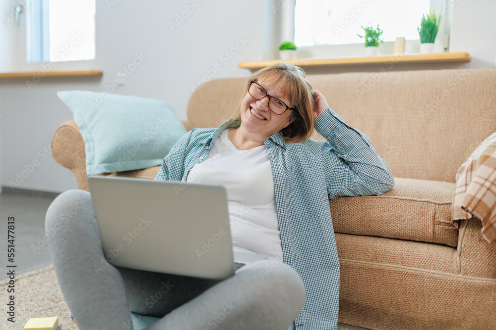 Happy blond businesswoman with laptop looking at you while sitting on the floor of living-room and surfing in the net Happy blond businesswoman with laptop looking at you while sitting on the floor of living-room and surfing in the net