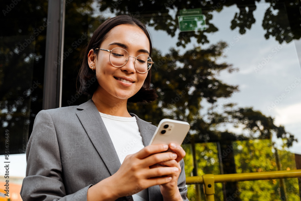 Young woman in glasses smiling and holding smart phone while standing near the bus