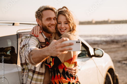 Happy white couple taking selfie photo by car while walking at seashore