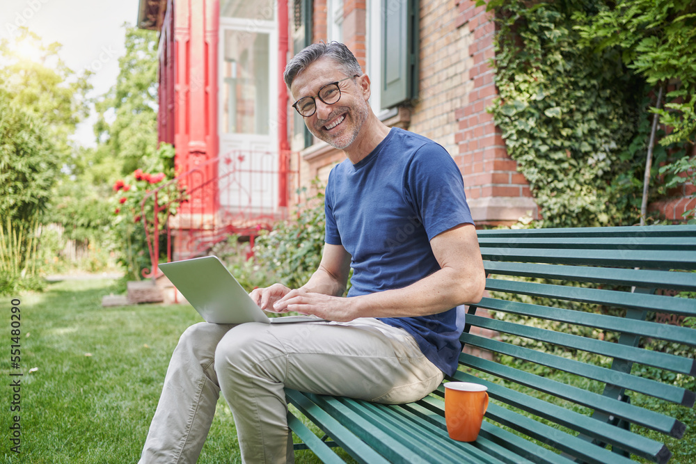 Happy businessman with laptop sitting by coffee cup on bench in back yard Happy businessman with laptop sitting by coffee cup on bench in back yard