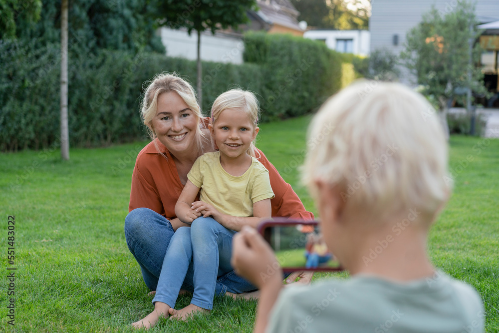 Boy photographing smiling mother and sister through smart phone in garden Boy photographing smiling mother and sister through smart phone in garden