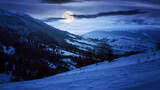 carpathian rural landscape in winter at night. snow covered hills in full moon light. scenery with krasna ridge in the distance. synevir village in the valley