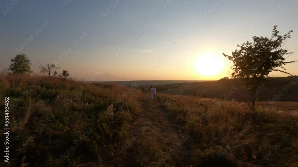 Cinematic FPV drone view of loving young man and woman hugging, kissing and holding hands on hilly meadow with green grass in bright sunlight, during beautiful summer sunset in nature, slow motion. Cinematic FPV drone view of loving young man and woman hugging, kissing and holding hands on hilly meadow with green grass in bright sunlight, during beautiful summer sunset in nature, slow motion.
