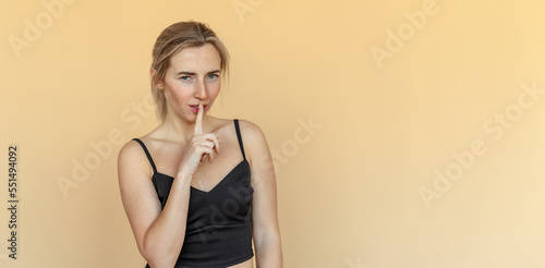 Cute young woman with freckles asking keep secret, take promise not tell or be quiet about it, standing over beige background. Shh shy sly people person modern concept. Copy space