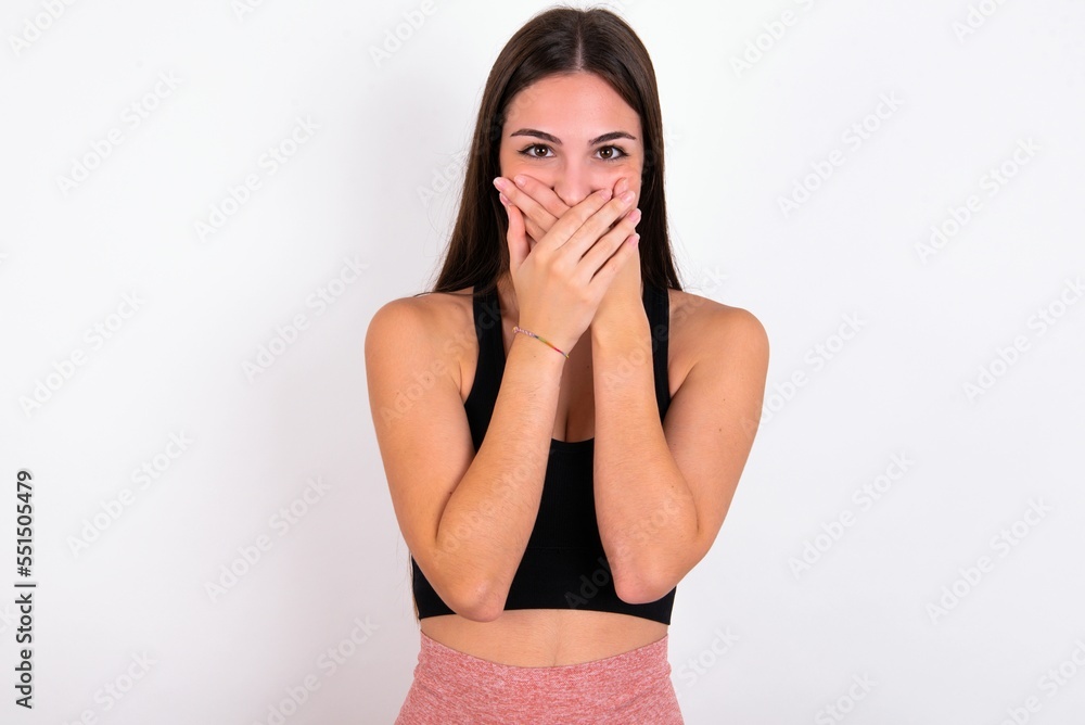 Fototapeta premium Upset Young caucasian woman wearing sportswear over white background , covering her mouth with both palms to prevent screaming sound, after seeing or hearing something bad.
