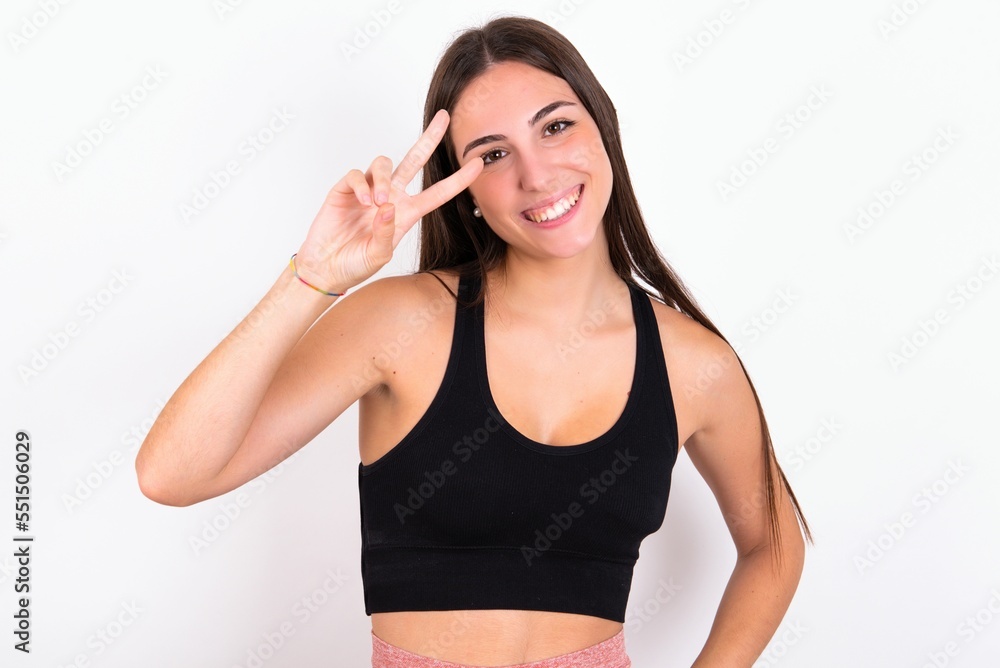 Young caucasian woman wearing sportswear over white background making v-sign near eyes. Leisure, coquettish, celebration, and flirt concept. Young caucasian woman wearing sportswear over white background making v-sign near eyes. Leisure, coquettish, celebration, and flirt concept.