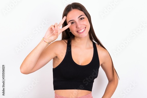 Young caucasian woman wearing sportswear over white background making v-sign near eyes. Leisure, coquettish, celebration, and flirt concept.