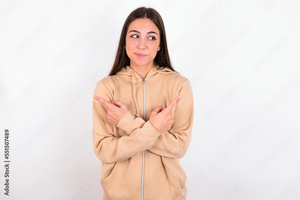 Young caucasian woman wearing sweatshirt over white background crosses arms and points at different sides hesitates between two items or variants. Needs help with decision Young caucasian woman wearing sweatshirt over white background crosses arms and points at different sides hesitates between two items or variants. Needs help with decision