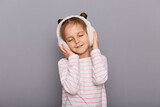 Portrait of relaxed cute charming little girl wearing striped shirt and fur headphones, keeps hands on her ears, posing with closed eyes, enjoying, standing isolated over gray background.