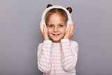 Portrait of little cute female kid standing in fur headphones and looking at camera with smile, expressing positive emotions, wearing striped shirt, being in good mood.