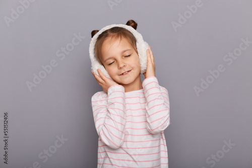 Portrait of relaxed cute charming little girl wearing striped shirt and fur headphones, keeps hands on her ears, posing with closed eyes, enjoying, standing isolated over gray background.