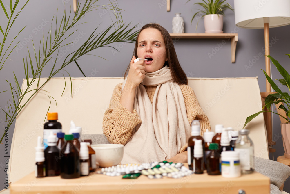 Φωτογραφία Portrait of ill sick young adult woman wearing warm jumper and scarf sitting on sofa in living room at home, having problems with health, has sore throat, using spray