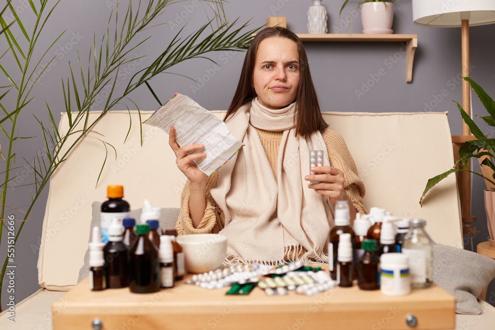 Horizontal shot of confused sad upset woman reads instructions before taking pills, wearing warm sweater and wrapped in scarf, looking at camera, feeling unwell. Horizontal shot of confused sad upset woman reads instructions before taking pills, wearing warm sweater and wrapped in scarf, looking at camera, feeling unwell.