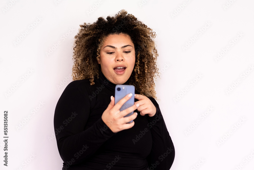 Surprised young woman looking at smartphone. Female African American model with curly hair and brown eyes in black turtleneck seeing shocking message or news at smartphone. Communication concept Surprised young woman looking at smartphone. Female African American model with curly hair and brown eyes in black turtleneck seeing shocking message or news at smartphone. Communication concept