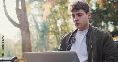 Caucasian young man sitting on bench in park on autumn day and working on laptop. outdoors. Male freelancer worker typing on computer in nature among trees in fall and resting. photo