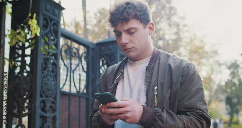 Caucasian young man walking the street with serious face and texting message on smartphone. Outdoor. Male strolling at park in fall and using mobile phone chat with strict face. Caucasian young man walking the street with serious face and texting message on smartphone. Outdoor. Male strolling at park in fall and using mobile phone chat with strict face.