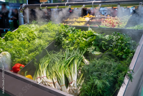 Sale of fresh green vegetables - lettuce salad, parsley, dill, green onions, herbs. Fresh herbs on display at grocery store under cooling water steam. Vegetables sprayed with mist water from nozzles