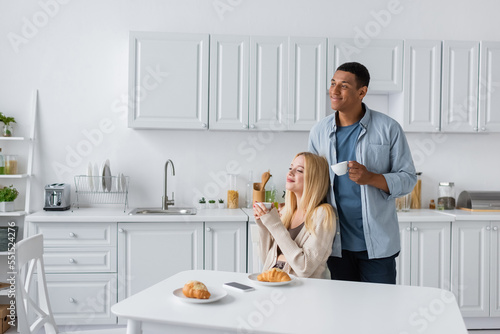 cheerful multiethnic couple with coffee cups looking away near delicious croissants in kitchen