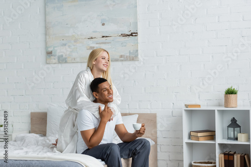 young woman in white silk robe hugging african american boyfriend sitting on bed with morning coffee photo