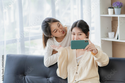 Young beautiful Asian LGBT lesbian happy couple sitting on sofa using smartphone in living room at home. LGBT lesbian couple living together