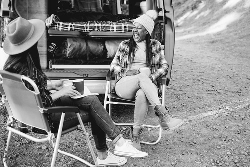 Multiracial women friends having fun camping with camper van while drinking coffee outdoor - Focus on african female face - Black and white editing