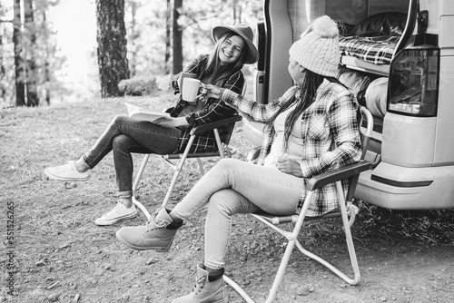 Multiracial women friends having fun cheering with coffee camping outside camper van - Focus on african female hand holding cup - Black and white editing