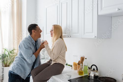 side view of laughing woman sitting on kitchen worktop and holding hands with happy african american boyfriend