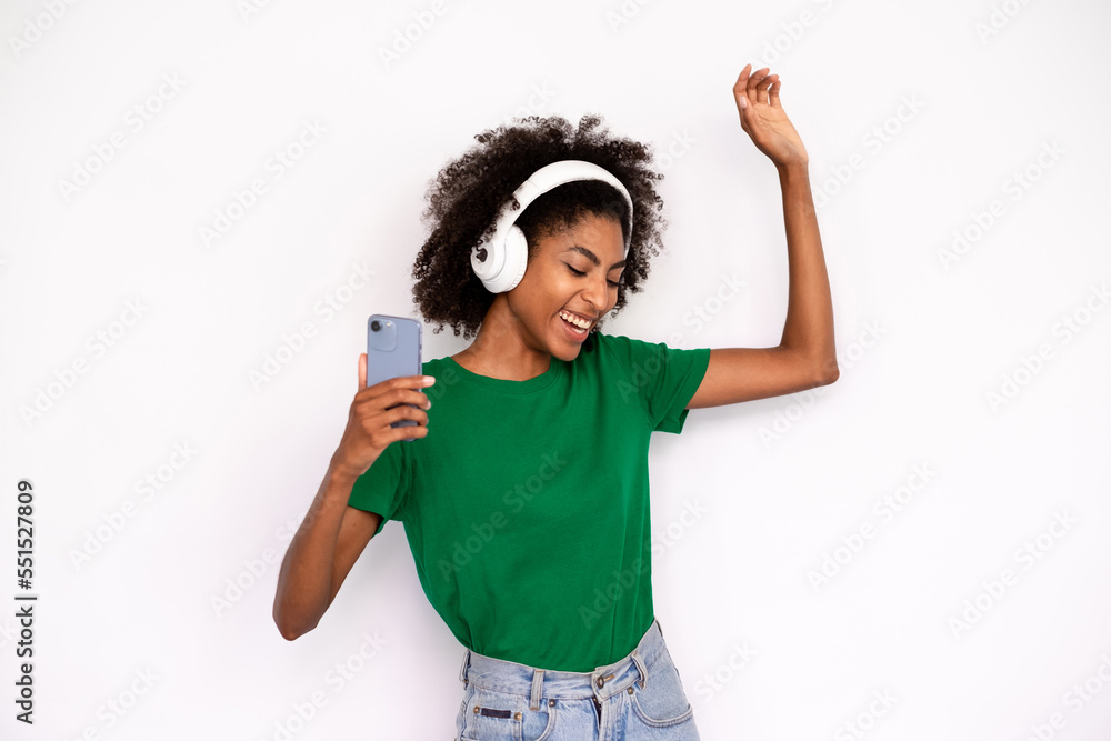 Portrait of excited young woman dancing in headphones over white background. African American lady wearing green T-shirt and jeans listening to phone player. Mobile technology concept Portrait of excited young woman dancing in headphones over white background. African American lady wearing green T-shirt and jeans listening to phone player. Mobile technology concept