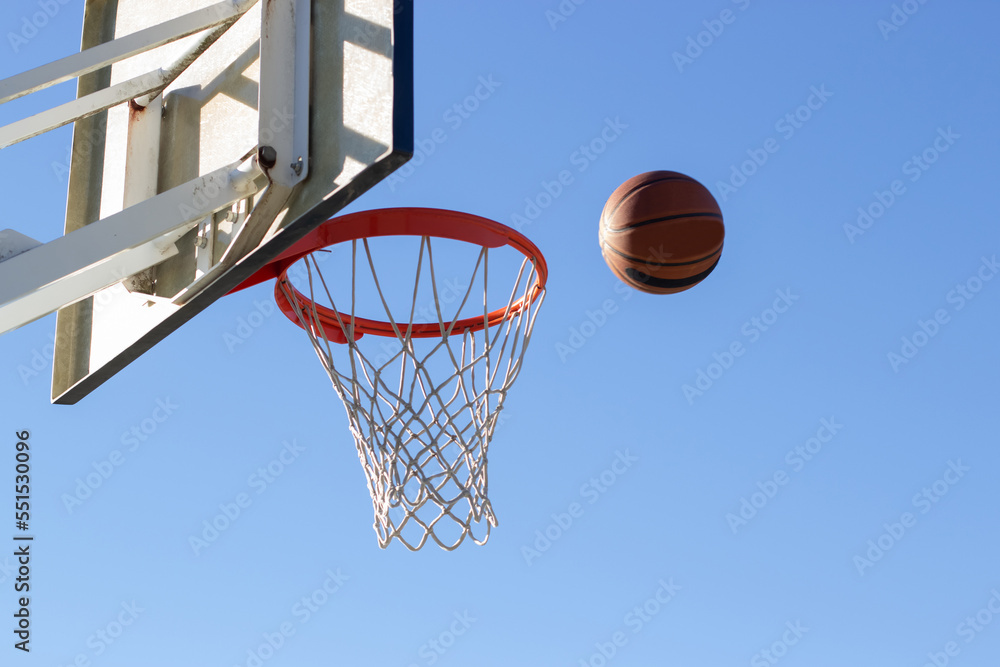 Low angle shot of basketball flying into hoop outside on sunny day. Ball thrown into basket with white backboard against blue cloudless sky. Achievement, sports concept
