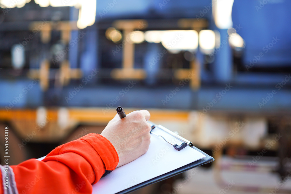 An engineer is writing on paper the list the maintenance detail with a heavy pumping machine as blurred background. Industrial expertise occupation working scene, selective focus at hand's part.