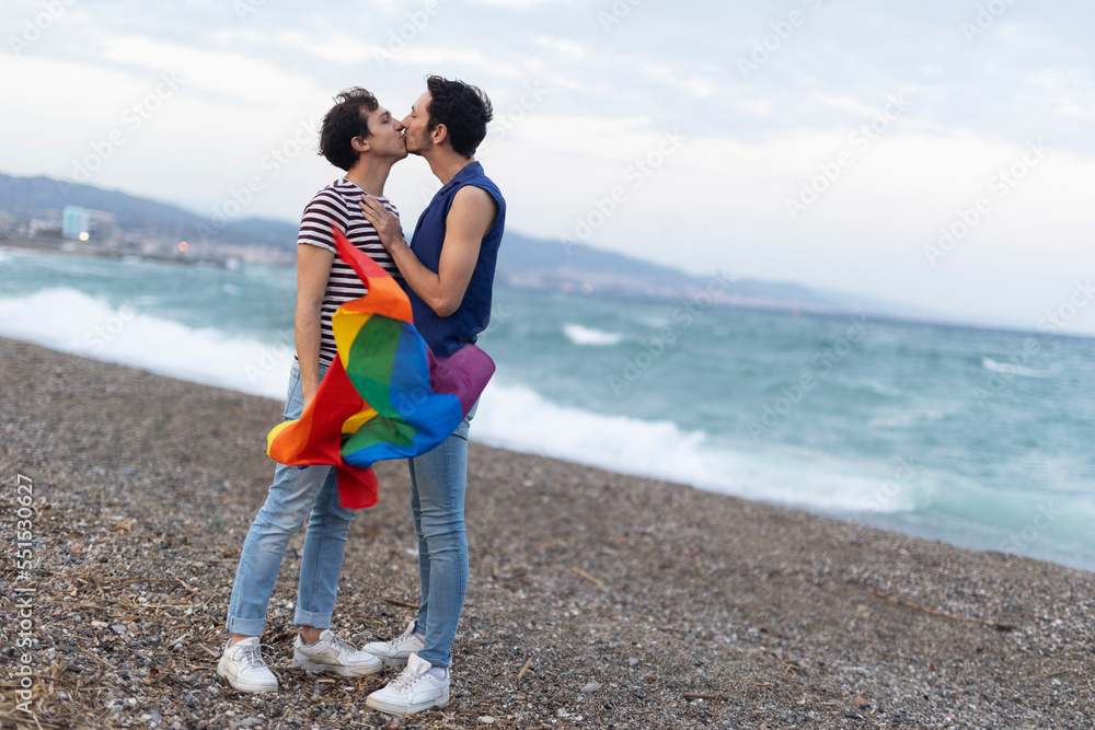 Beautiful gay young couple embraces and holds a rainbow flag. Happy couple enjoy at the beach...
