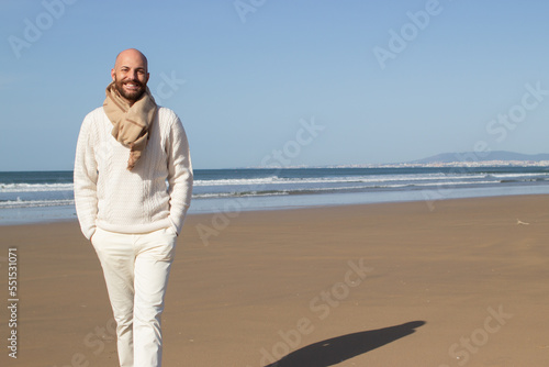 Smiling handsome man walking alone on beach. Happy bald tourist in scarf holding hands in pockets of white jeans. Resting at sea concept © KAMPUS