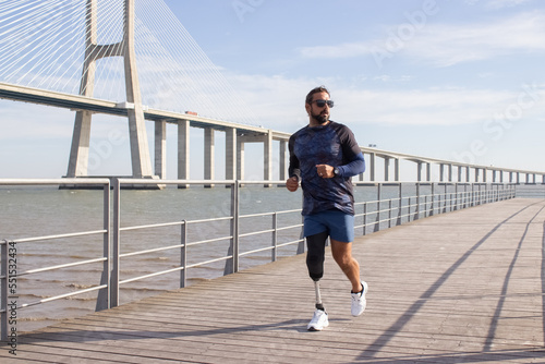 Handsome man with mechanical leg running on embankment. Caucasian man in sportive clothes jogging on summer day. Sport, leisure, disability concept