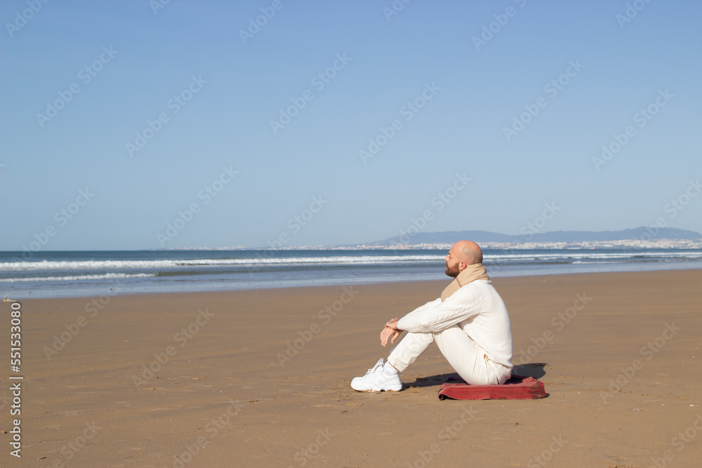 Lonely middle aged man in scarf sitting on beach. Bald man in white shoes looking at ocean. Inspiration and planning concept