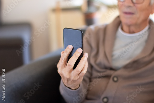 Senior man calling online via smartphone app. Closeup of aged man in cardigan sitting in armchair and using gadget at home. Technology concept