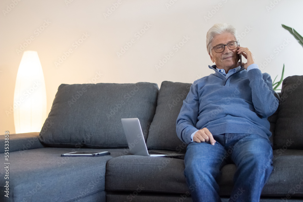 Content senior businessman working at home. Confident elderly man in blue sweater sitting on sofa with devices and talking on phone. Technology concept
