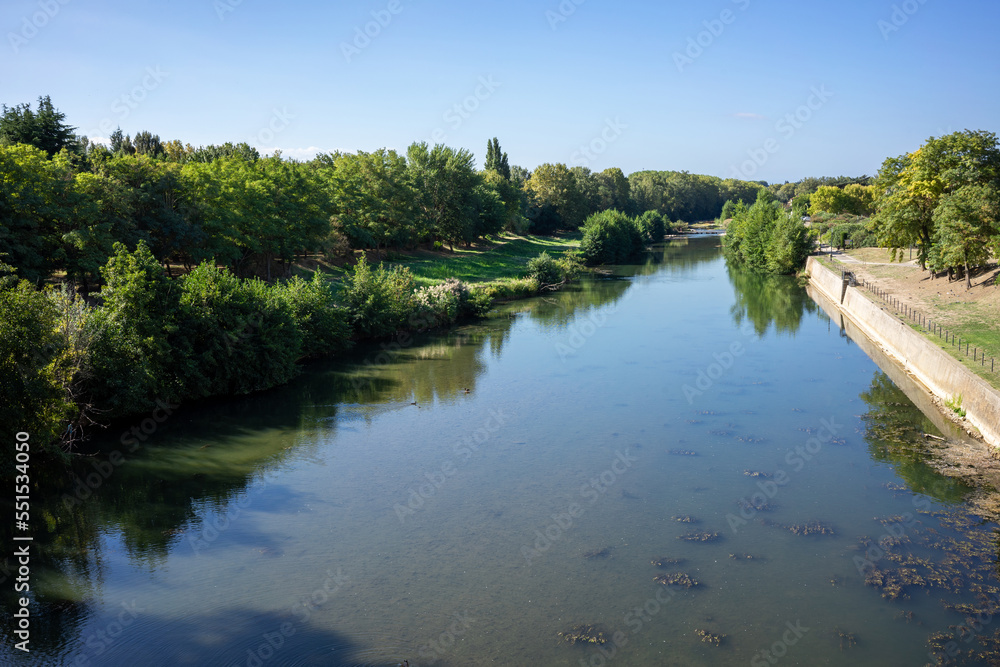 View of the Aude river in the French village of Carcassonne. It's a summer day with blue sky and green trees and bushes on the shore.