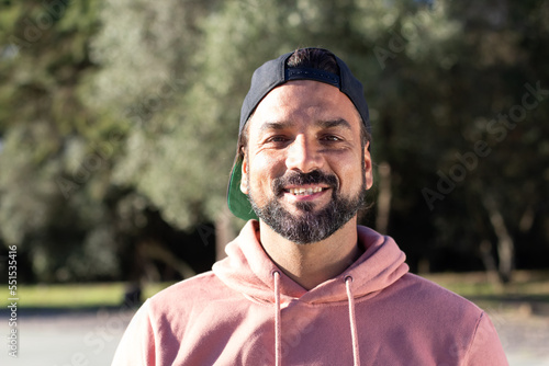 Happy Hispanic man smiling while spending time in park on sunny day. Portrait of friendly bearded guy in cap and pink hoodie posing outdoors, looking at camera. Lifestyle, happiness concept photo