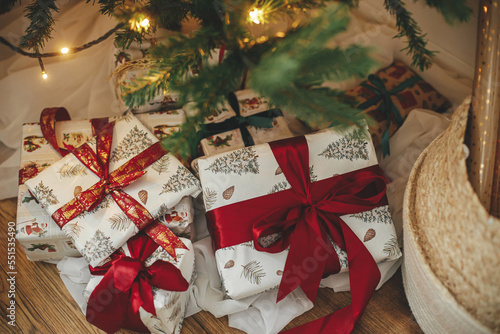 Stylish christmas gifts at christmas tree with golden lights bokeh. Wrapped christmas presents with red ribbon under decorated tree in room. Atmospheric eve. Holiday banner. Merry Christmas!