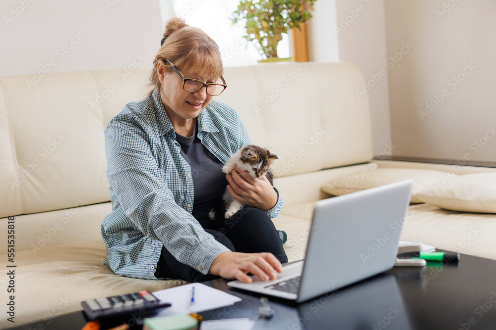 Mature woman in blue shirt sitting with a cat on her lap at the wooden table at home with laptop and notebook, working or shopping online.
Mature woman in blue shirt sitting with a cat on her lap at the wooden table at home with laptop and notebook, working or shopping online.