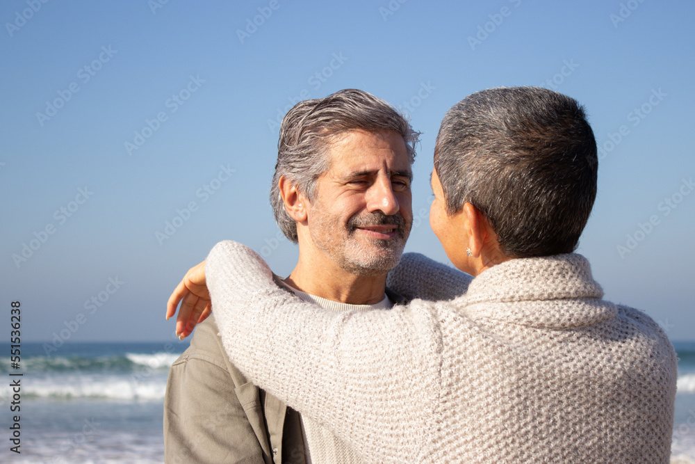 Handsome senior man embracing his wife and smiling while short-haired woman laying arms on mans shoulder. Middle-aged couple enjoying day at seashore. Sea background. Relationship, romance concept Handsome senior man embracing his wife and smiling while short-haired woman laying arms on mans shoulder. Middle-aged couple enjoying day at seashore. Sea background. Relationship, romance concept